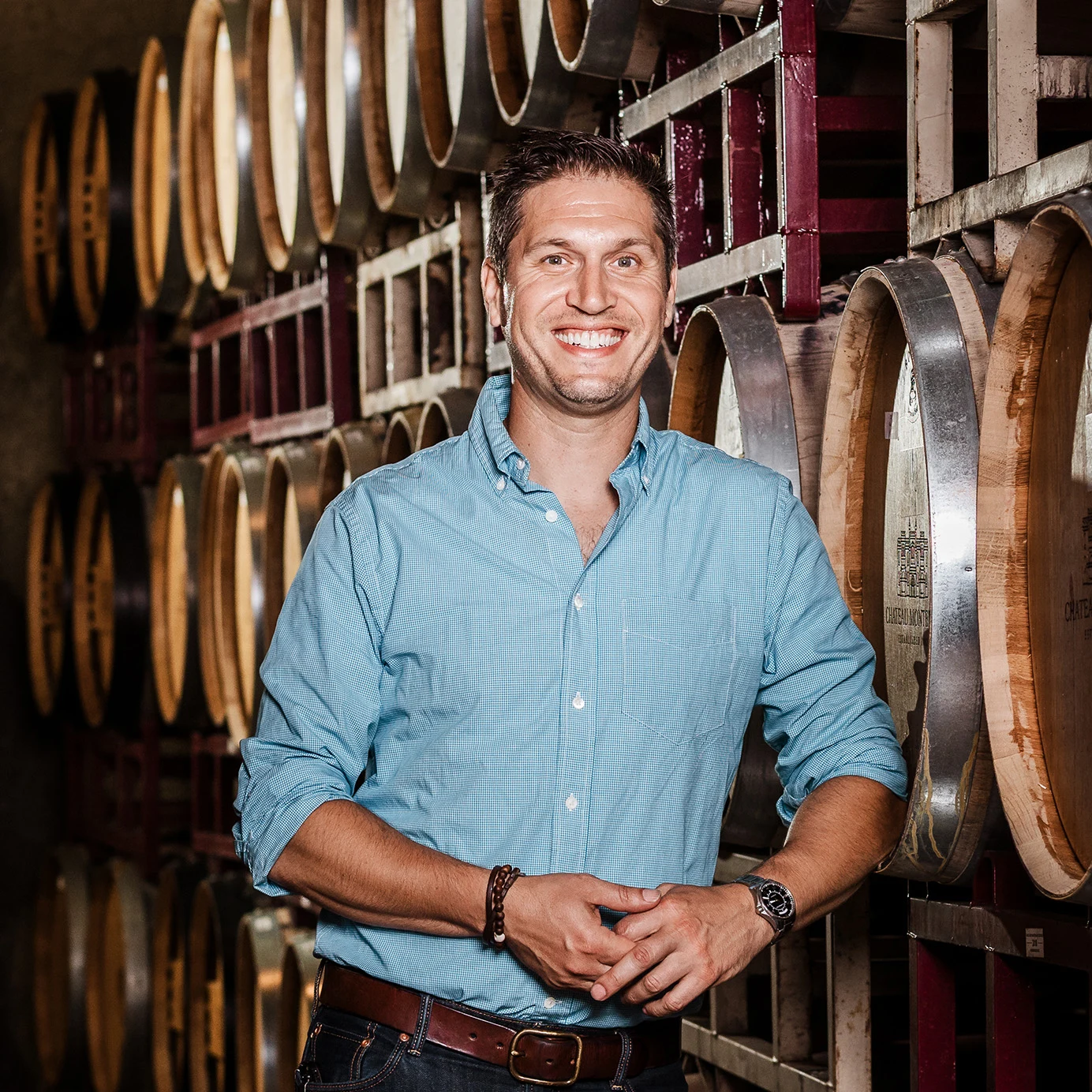 Professional portrait of a man in a light blue button-down shirt standing in a wine barrel room.