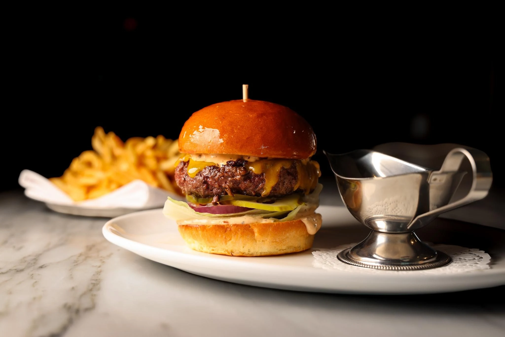 A gourmet burger with melted cheese on a brioche bun, served with fries and a sauce boat on a white plate