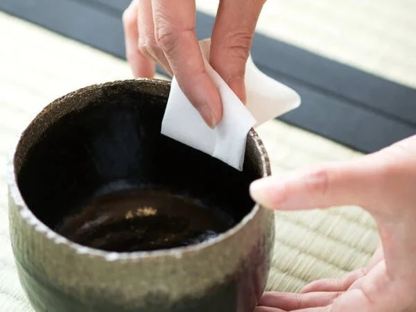 A close-up of hands performing a tea ceremony ritual, with one hand holding a white folded cloth (chakin) wiping the rim of a dark ceramic tea bowl.