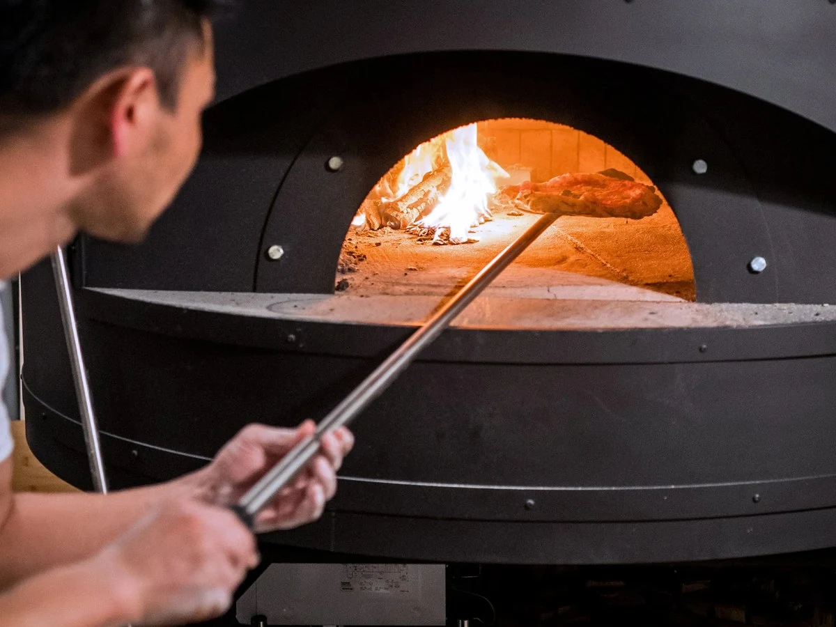pizza studio tamaki: A chef tends to a wood-fired pizza oven, carefully rotating a pizza with a long peel.