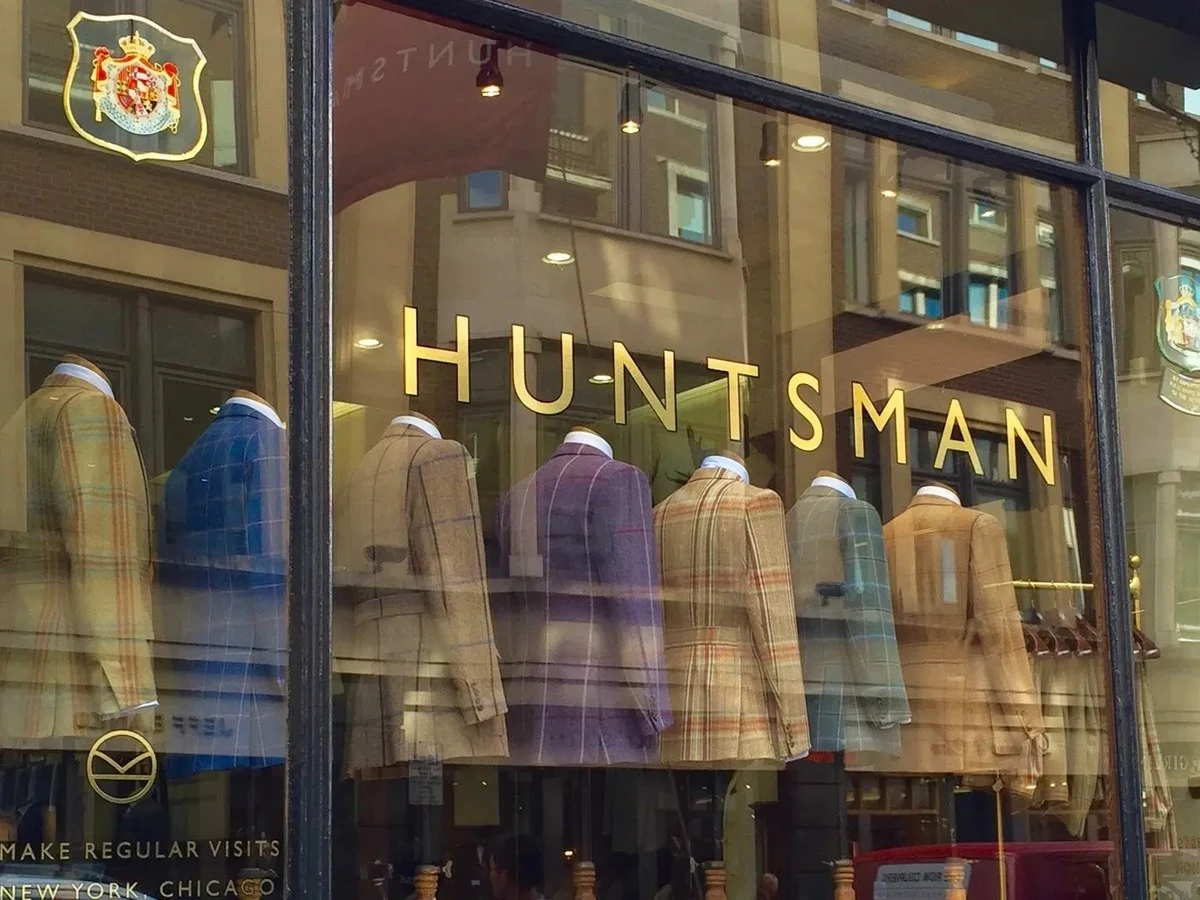 The exterior shopfront of Huntsman on Savile Row, featuring a large display window with multiple tweed jacket mannequins and the gold 'HUNTSMAN'