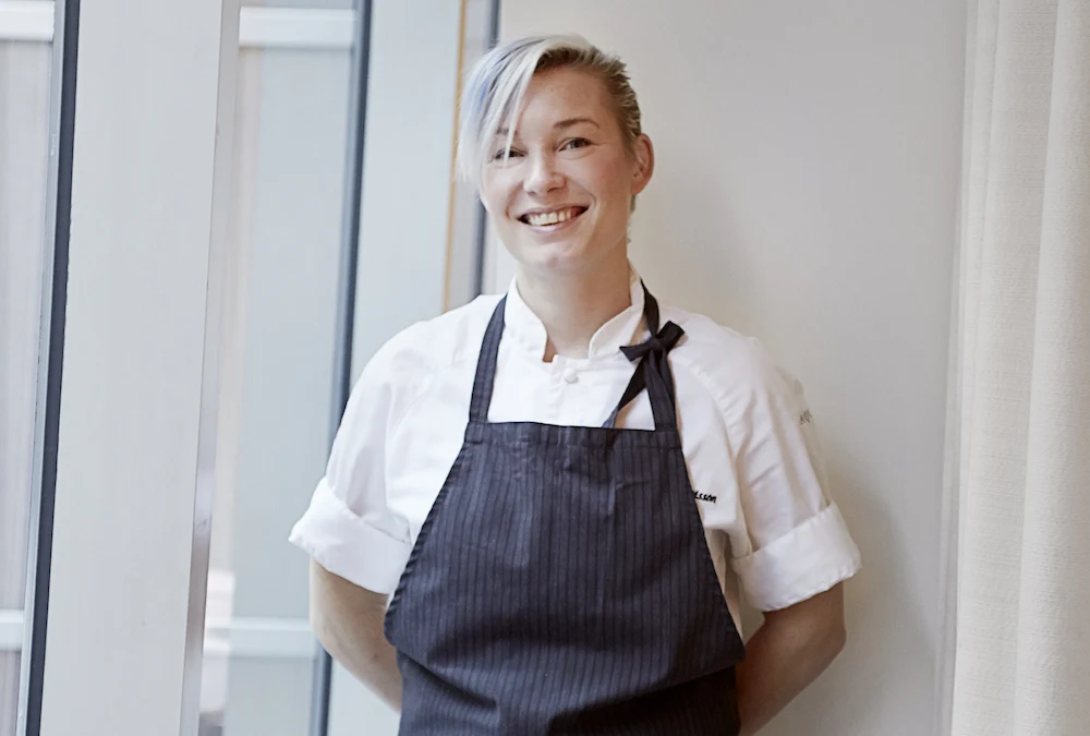 A chef in white jacket and dark striped apron stands in a bright, modern interior; the source page explicitly attests this is Emma Bengtsson via alt-t