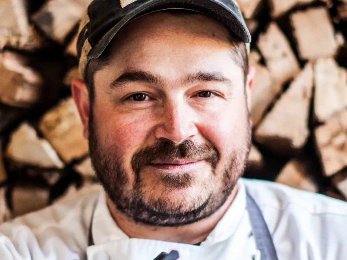 Chef in white coat and gray apron standing before stacked firewood.