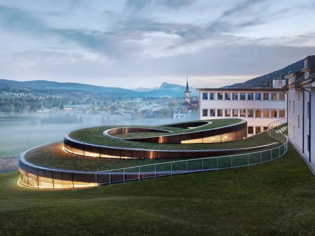 A modern spiral-shaped building with a green roof, illuminated from within, connects to a traditional white multi-story hotel.