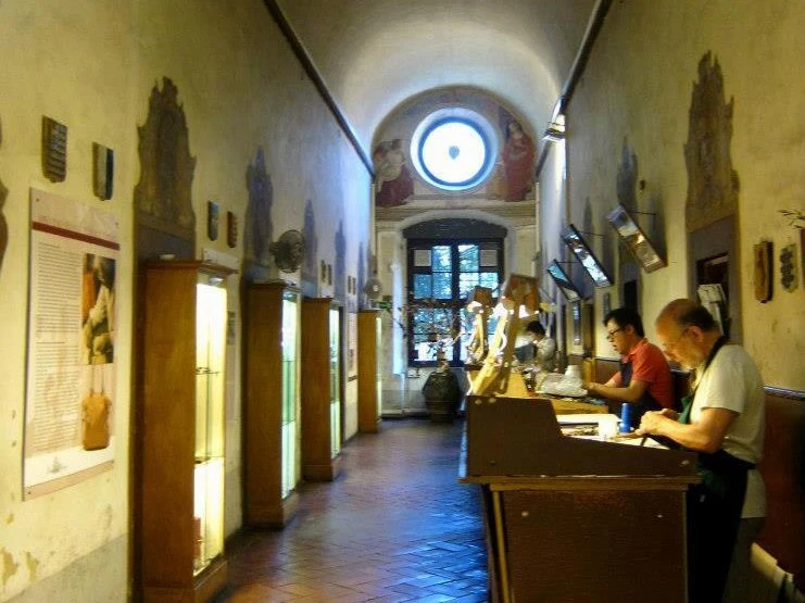A long, vaulted corridor with terracotta floors and frescoed walls, where two men work at a wooden bench in a leather workshop.