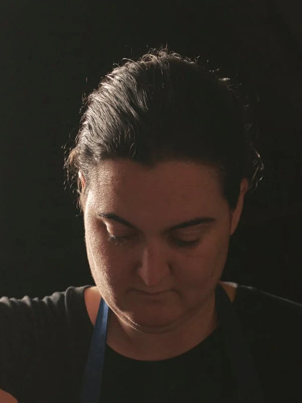 Portrait photograph of a person in chef's clothing, shown in profile with eyes downcast against a dark background.