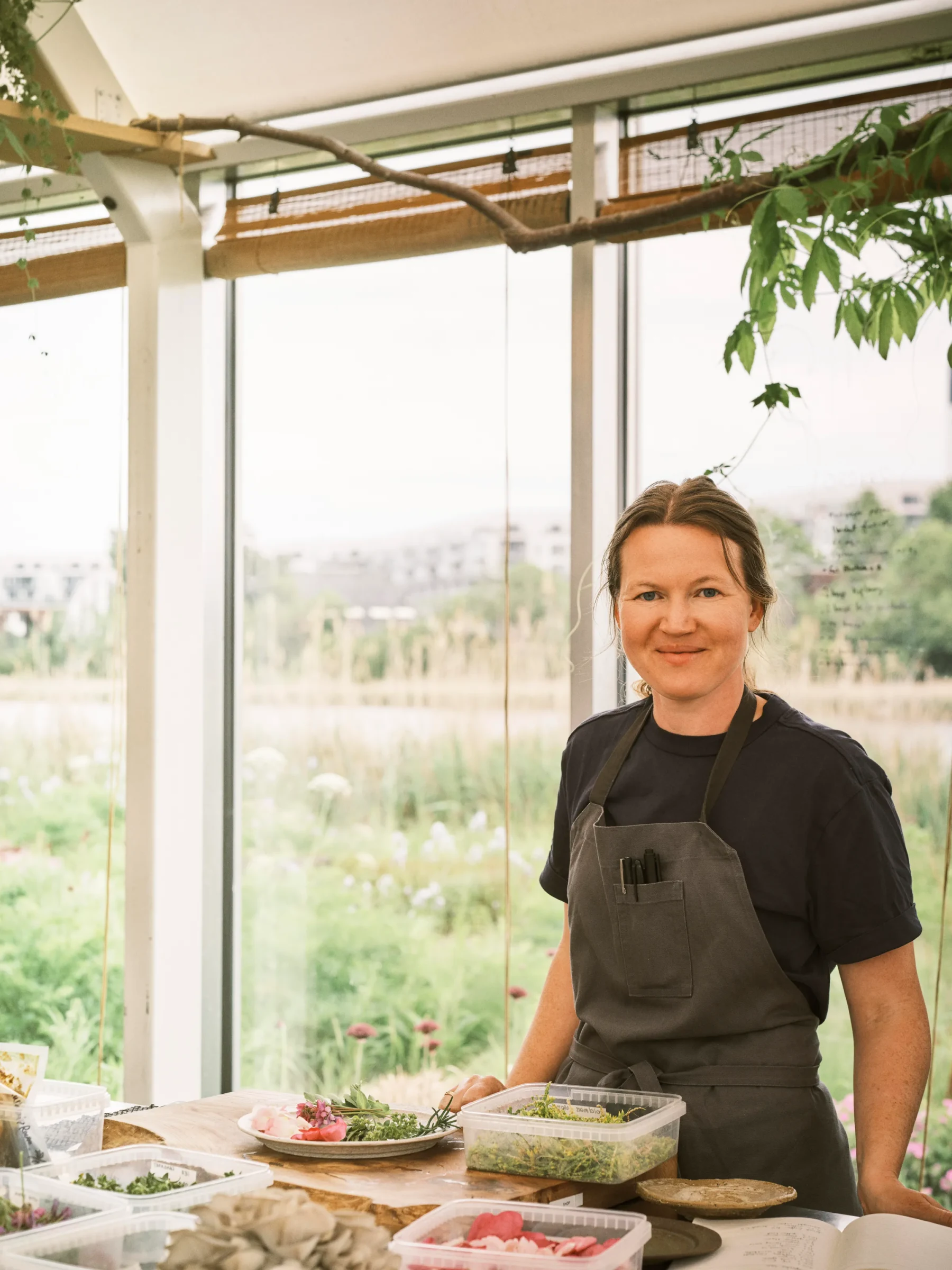 Mette Brink Søberg, a chef, smiles while standing behind a wooden table with various ingredients in clear containers.