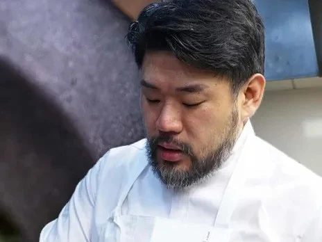A chef with dark hair and a beard wearing a white chef's coat looks down while working.