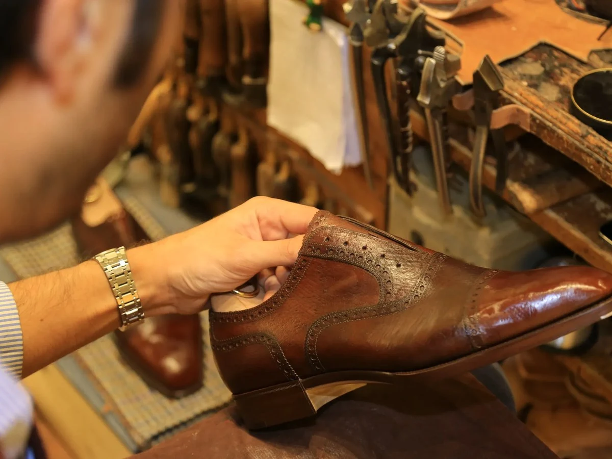 A person wearing a gold bracelet inspects a brown leather dress shoe with decorative stitching details on a work surface.