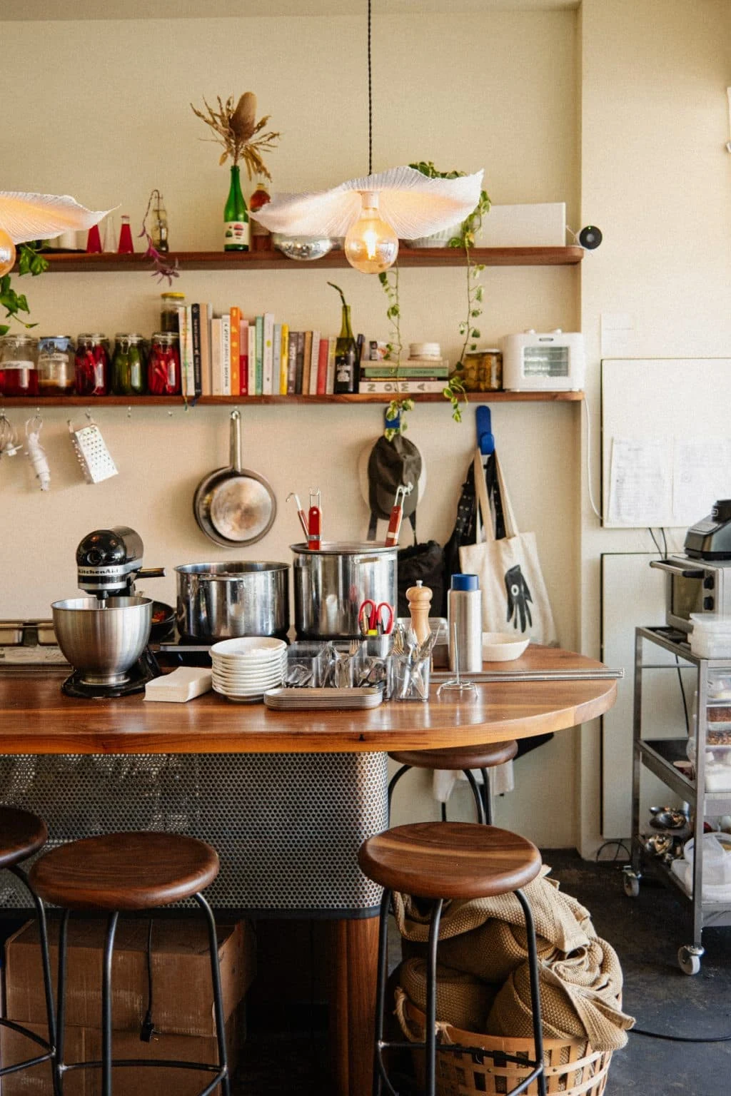 A kitchen with three wooden floating shelves mounted on a beige wall, stocked with cookbooks, jars, and decorative items, with a round wooden counter below featuring two wooden stools and various cooking equipment and utensils.