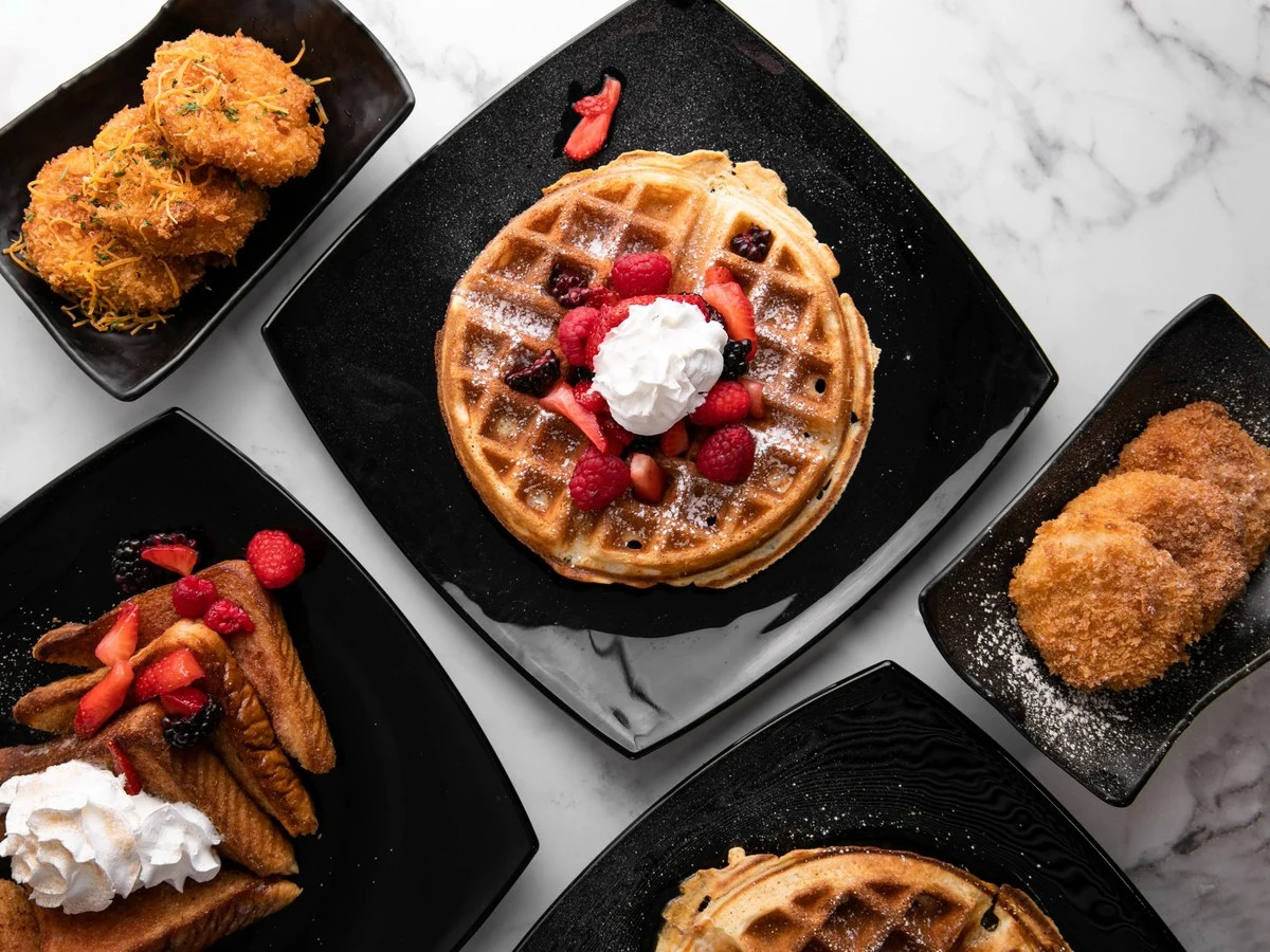Multiple plates of Mexican-inspired dishes including waffles with berries and whipped cream, fried items dusted with sugar, and breaded items, present