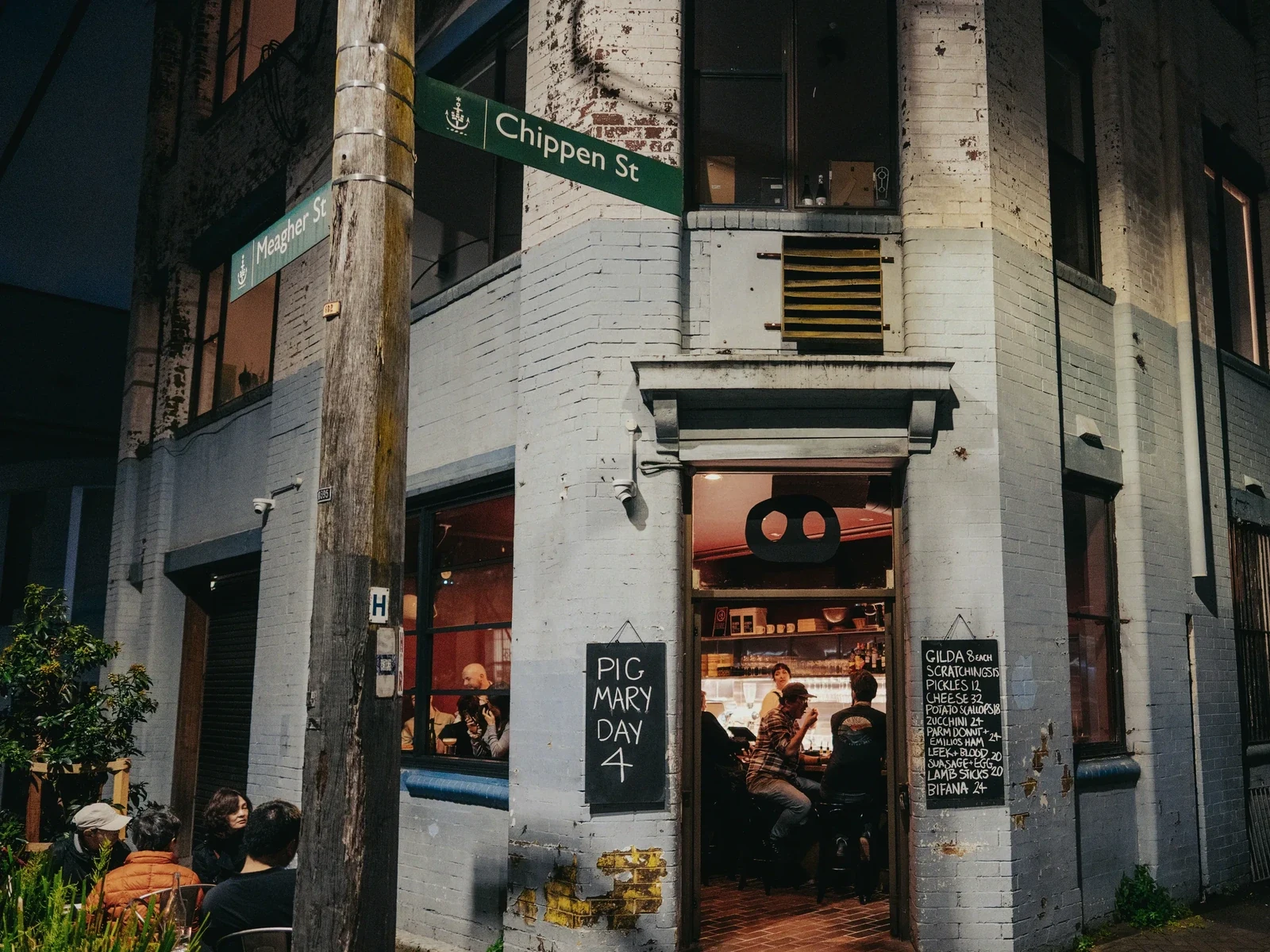 A corner storefront with white brick facade at night, green street signs reading Chippen St and Meagher St, red door entrance, blackboard menu visible, and people dining inside.