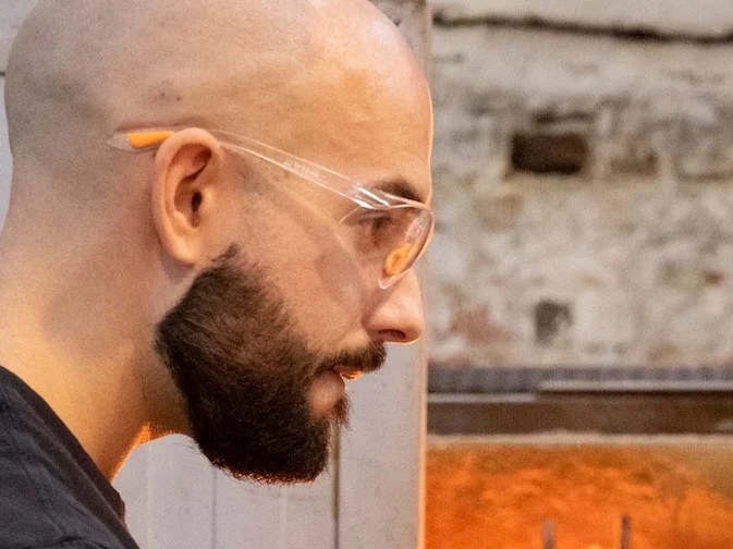 A bald, bearded man in safety glasses and protective gloves holds a blowpipe with glowing molten glass in front of a furnace, observed by a woman in