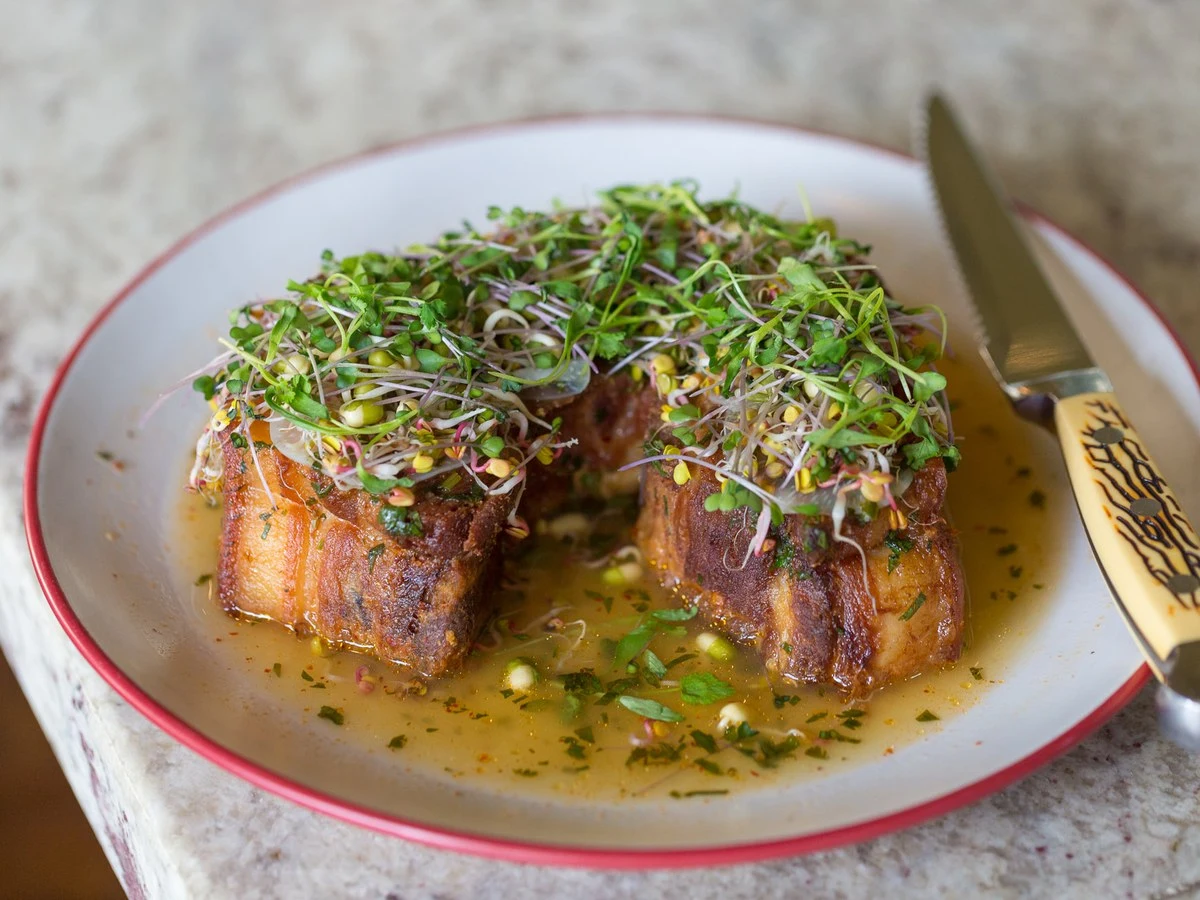 Two pieces of crispy pork belly, topped with a vibrant mix of green and purple microgreens, sit in a shallow pool of sauce on a white plate.