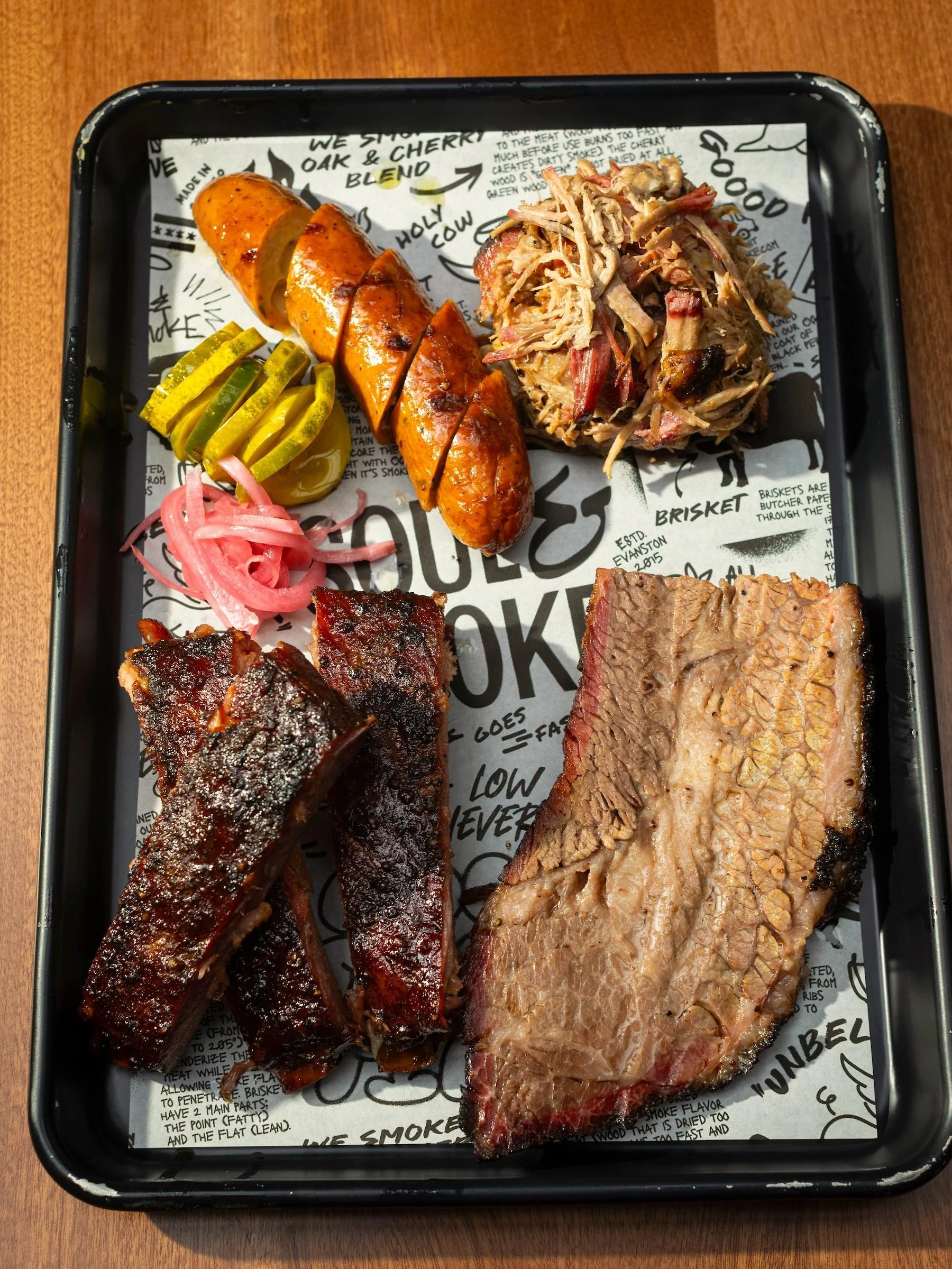 A black tray lined with printed paper displays various smoked and grilled meats, pickled vegetables, and barbecue accompaniments arranged for serving.