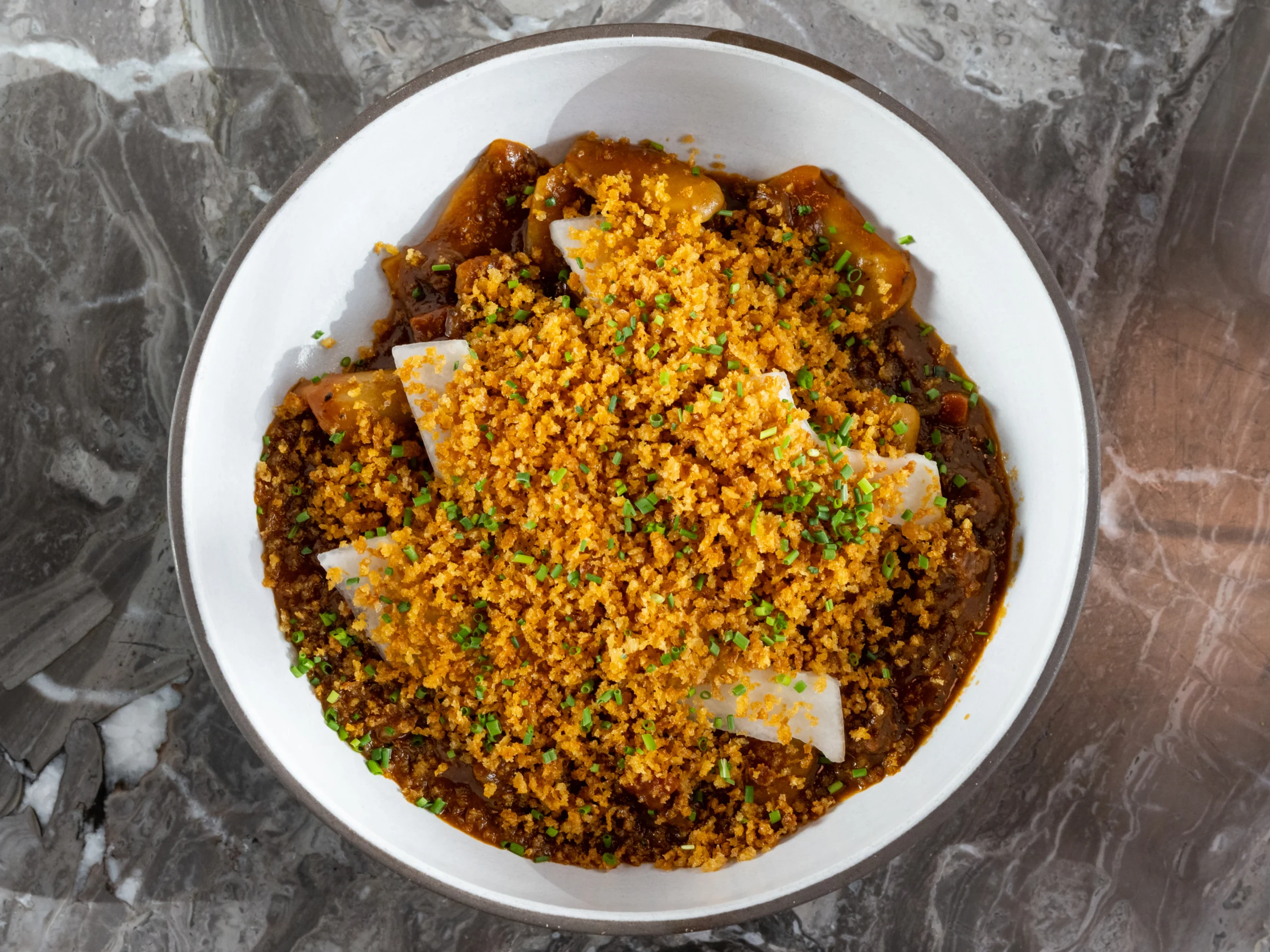 Overhead view of a white bowl containing braised meat covered with a crispy golden breadcrumb topping, green onion garnish, and sesame seeds on a gray marble surface.
