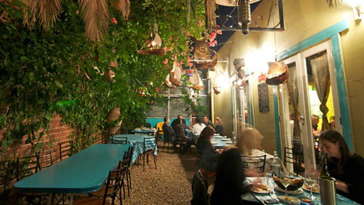 An outdoor courtyard dining area with turquoise tables, string lights, bougainvillea, and lush greenery; the source page (Time Out Melbourne) explicit