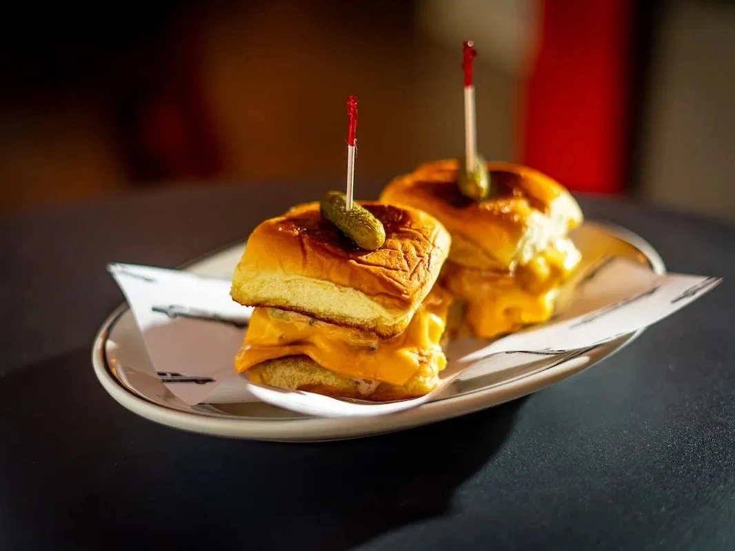 Two cheeseburger sliders with melted cheese and pickles on brioche buns, held by red toothpicks, on a white plate in a dimly lit bar.
