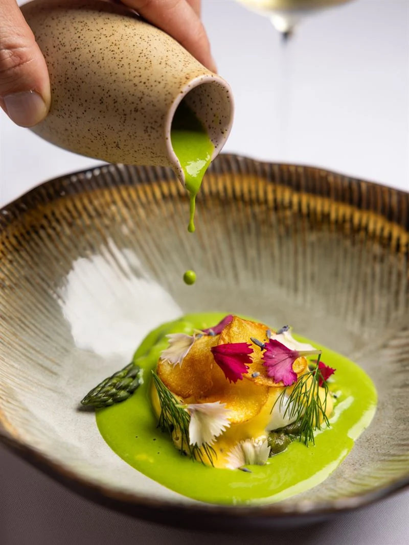A refined French dish with asparagus, edible flowers, dill, and a vibrant green herb sauce being poured tableside from a speckled ceramic vessel; the 