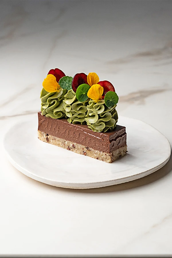 A plated dessert slice with a nut-based crust, chocolate layer, and matcha green tea piped topping decorated with edible flowers, published on So Good