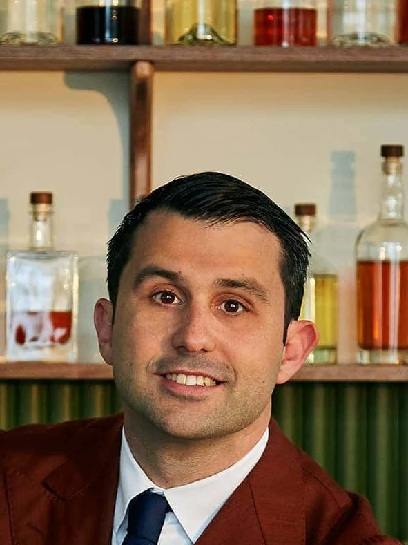 A professional portrait of Giorgio Bargiani, a man in a rust-colored suit, smiling behind a bar with empty cocktail glasses and bottles.
