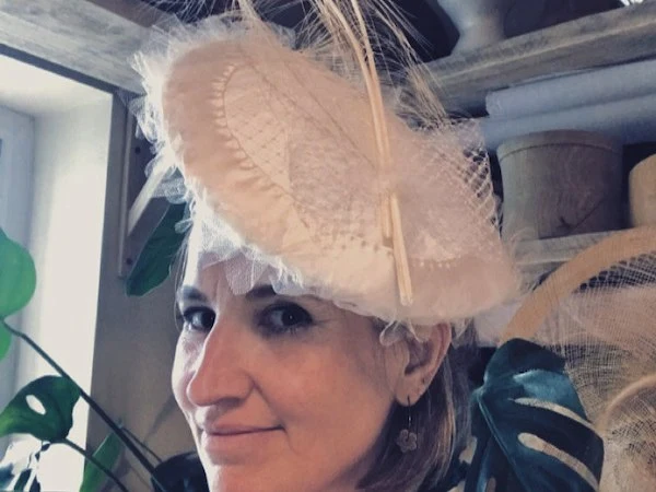 A woman, Chloë Scrivener, wears an elaborate cream fascinator hat with tulle and feather details, in a millinery studio with hat forms and plants.