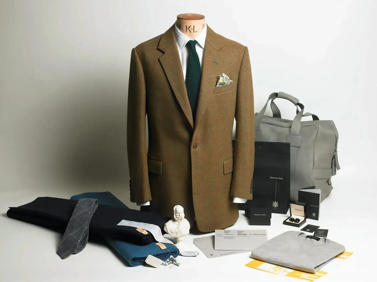 A tailoring mannequin wearing a brown tweed sport coat with green knit tie and floral pocket square, surrounded by folded fabrics, accessories,