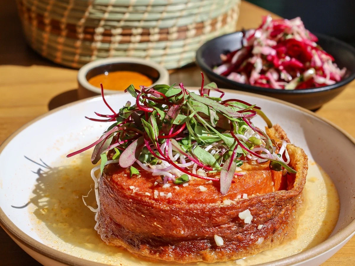 A close-up of a dish from Broken Spanish Comedor, featuring a cylindrical, crispy-edged item in a shallow pool of sauce, topped with fresh.