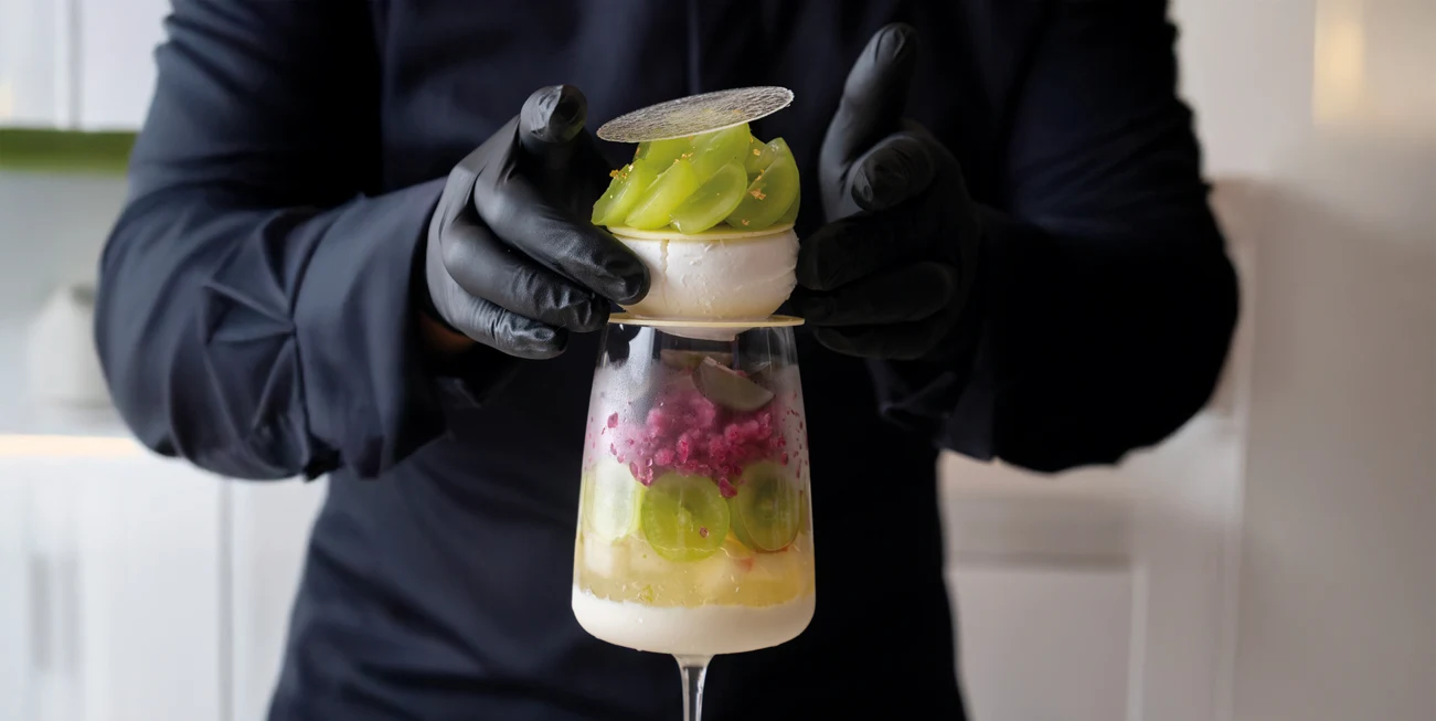 A person wearing black gloves presents an elaborate layered dessert in a glass with green grapes, pink granita, cream layers, and decorative garnishes