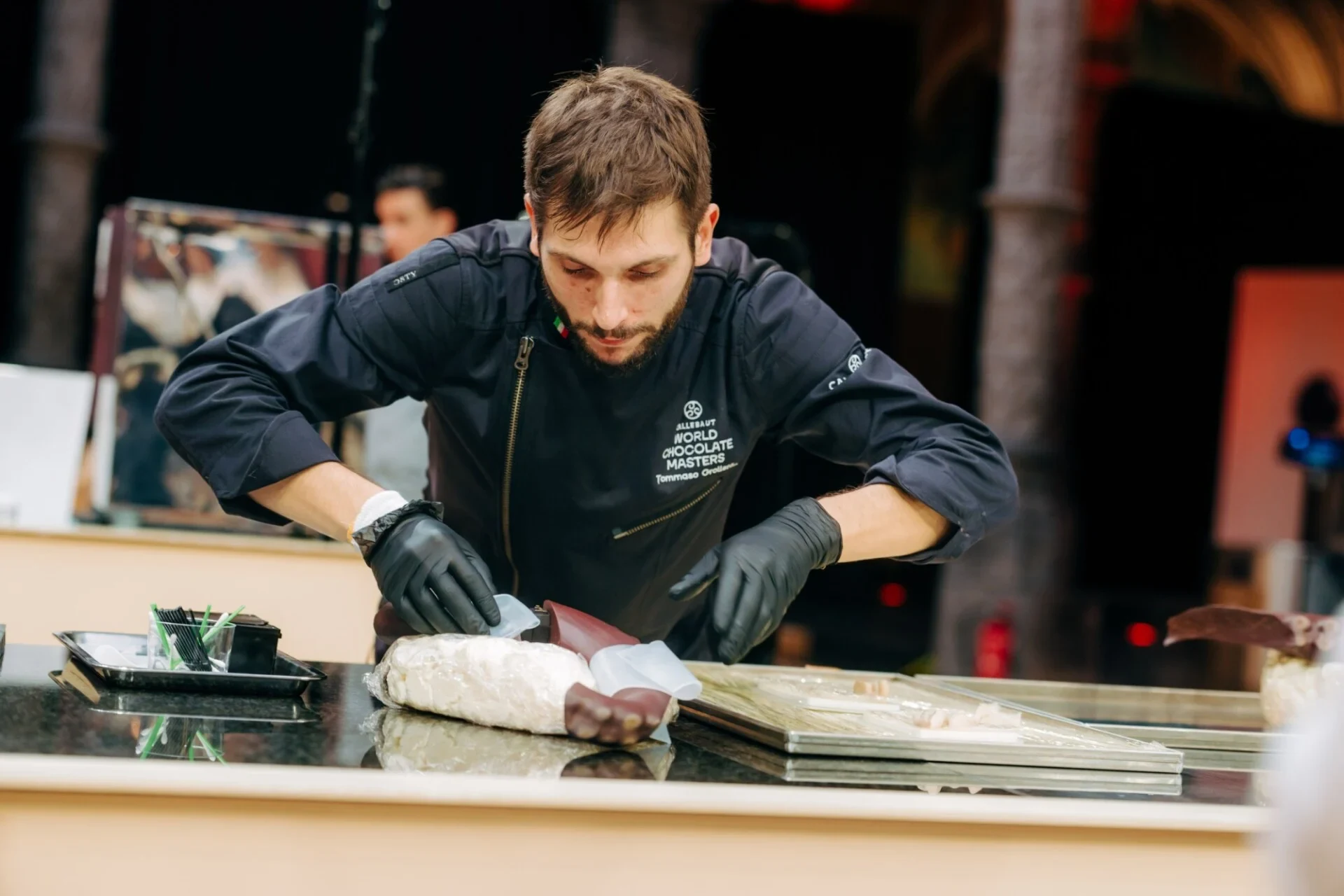 A chocolatier in black chef's jacket with 'World Chocolate Masters' branding works at a competition station with chocolate materials and tools; the so