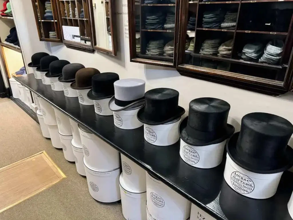 A row of black and brown bowler hats and top hats are displayed on white hat boxes, with glass display cases of flat caps on the wall.
