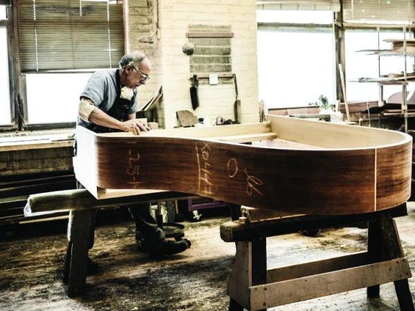 A craftsman meticulously shapes the wooden frame of a grand piano in a sunlit workshop filled with tools and wood.
