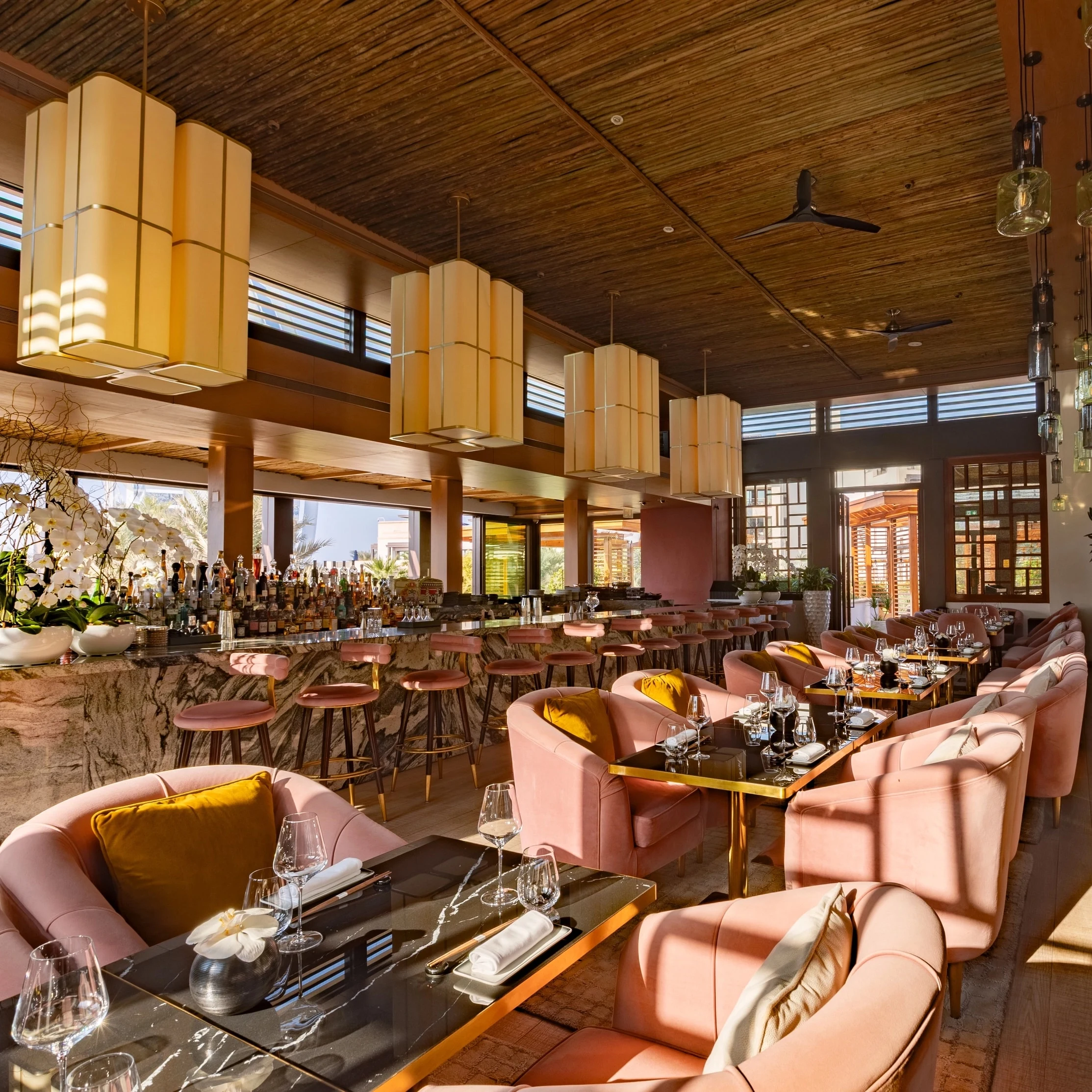 Upscale restaurant dining room interior with pink velvet seating, dark wood tables, large geometric pendant lights, and a wood-paneled ceiling; the so
