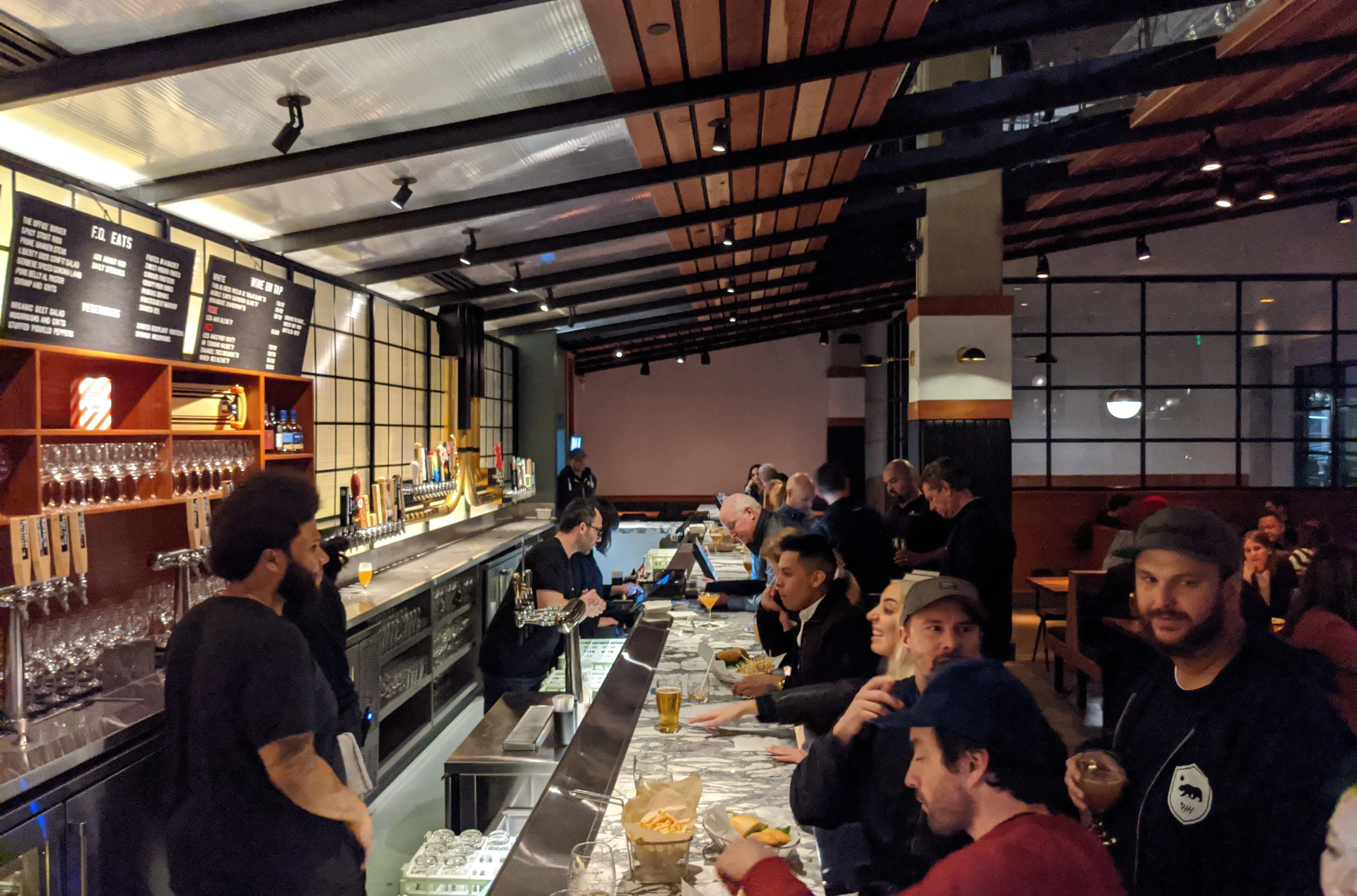 Modern gastropub interior with long bar counter, patrons seated along it, exposed beam ceiling, industrial-modern design elements, and menu boards; so