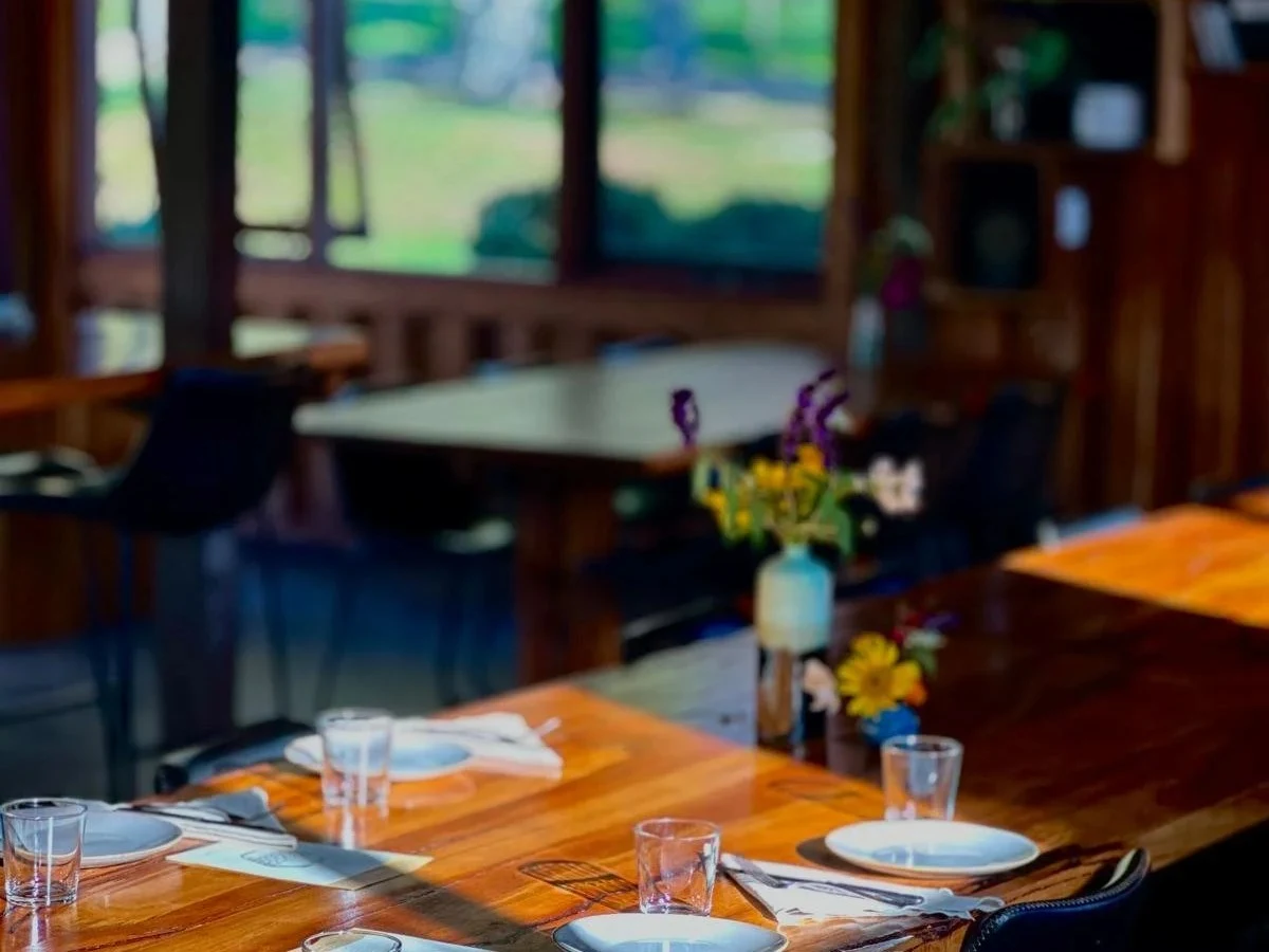 A warmly lit farm-to-table restaurant dining room interior featuring a long live-edge timber communal table set with white plates, glassware.