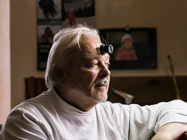 An older white-haired man wearing a white watchmaker's coat and loupe headset sits at a traditional wooden watchmaker's bench surrounded by precision