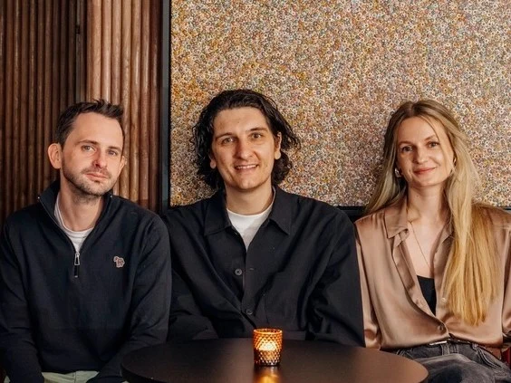 Three people sitting at a table in front of patterned wallpaper, with a small glass visible on the table in front of them.