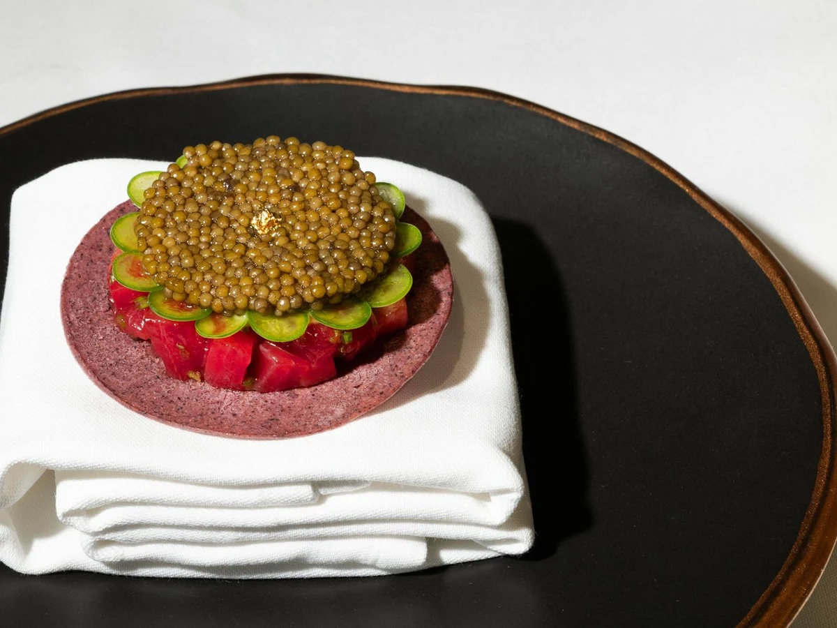 An elegantly plated fine-dining dish featuring caviar atop thinly sliced vegetables arranged in a flower pattern on a purple base, presented on a folded white napkin.