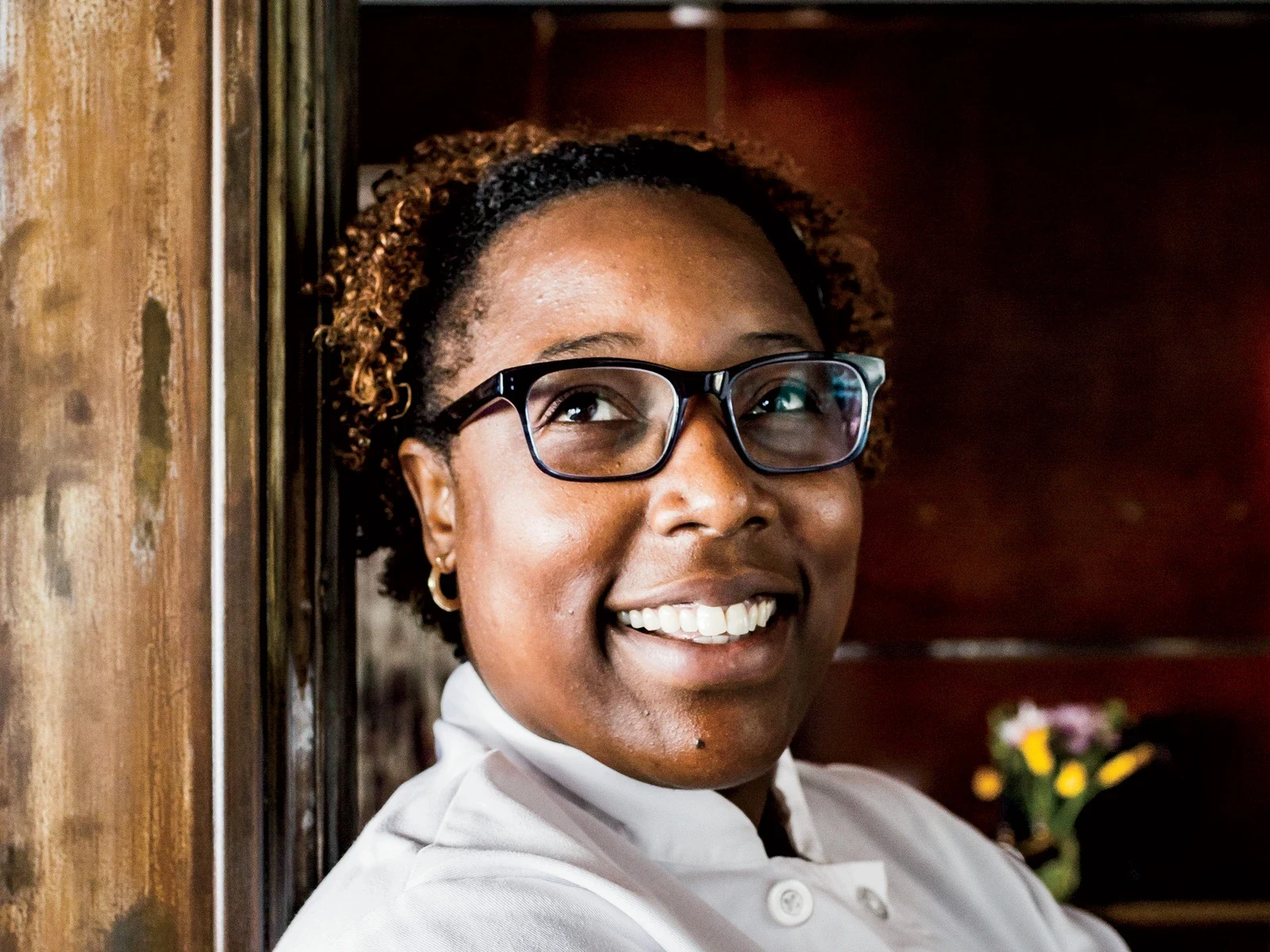 A smiling chef in glasses and a white coat stands beside a wooden column under hanging lights.