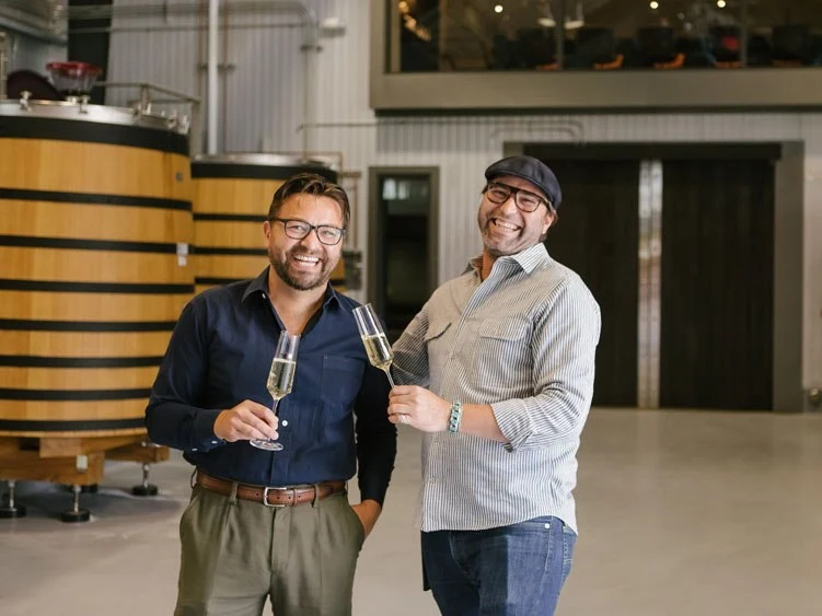 Two men in business casual attire holding champagne glasses in a winery production facility with large wooden barrels visible in the background; the s