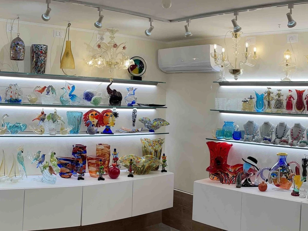 A brightly lit retail showroom displaying dozens of colorful Murano glass art objects, including figurines, vases, animals, chandeliers, and jewelry.
