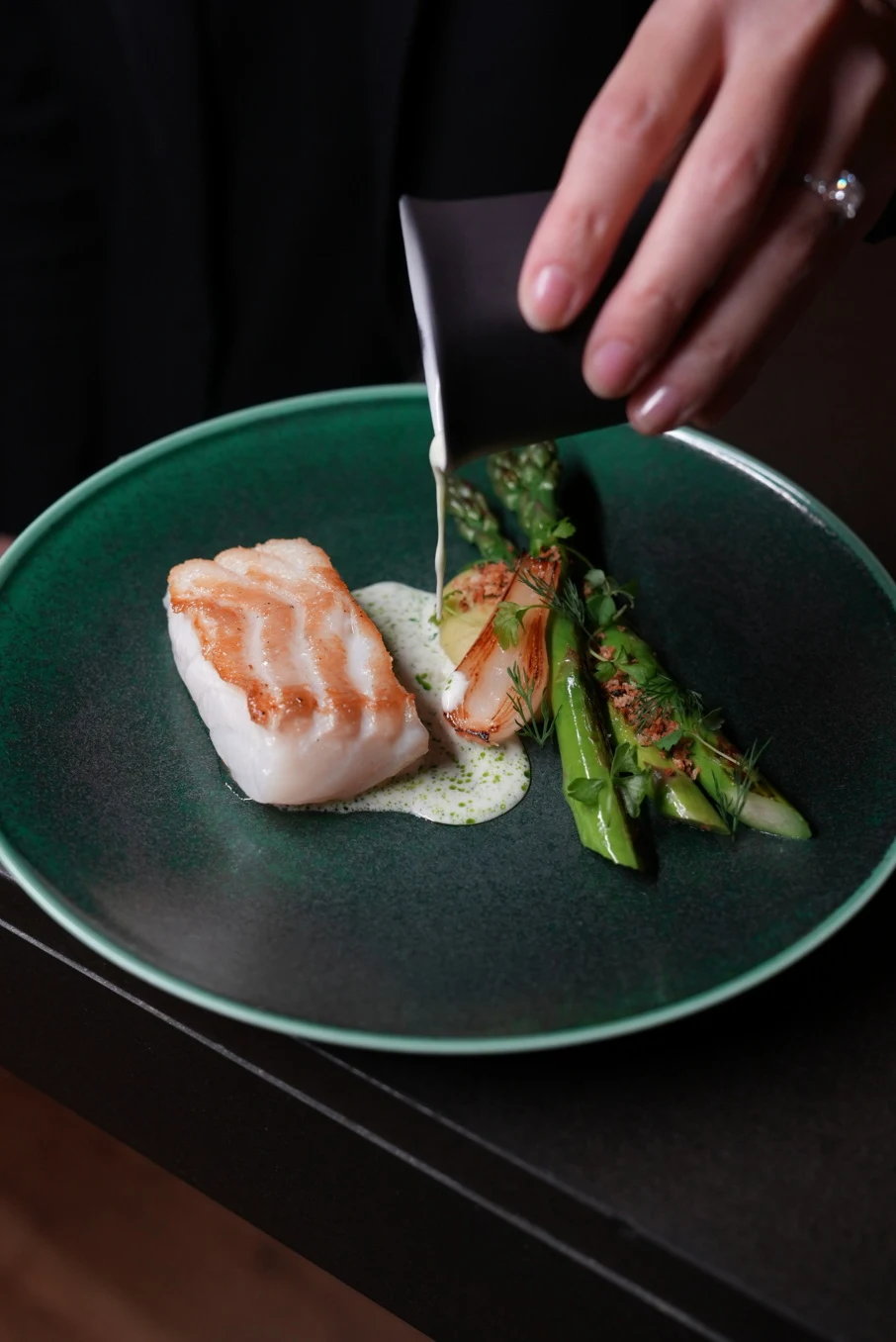 A plated seafood dish (lobster with braised endive, smoked yogurt, and chicories) on a dark green plate, photographed in editorial style with a hand p