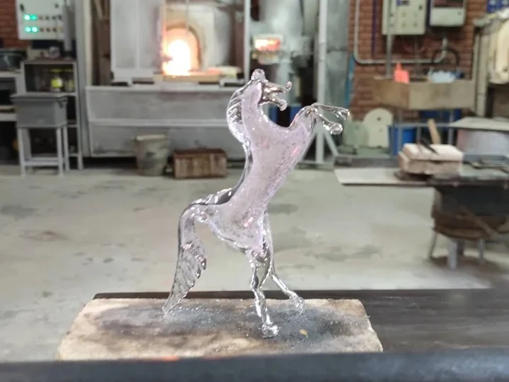 A clear glass horse figurine rests in a murano glass workshop, with a glowing furnace in the background.