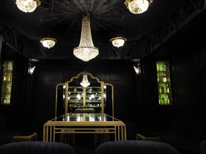 A dramatically lit, dark and moody speakeasy-style bar interior featuring crystal chandeliers, black paneled walls, gold-accented bar furniture.