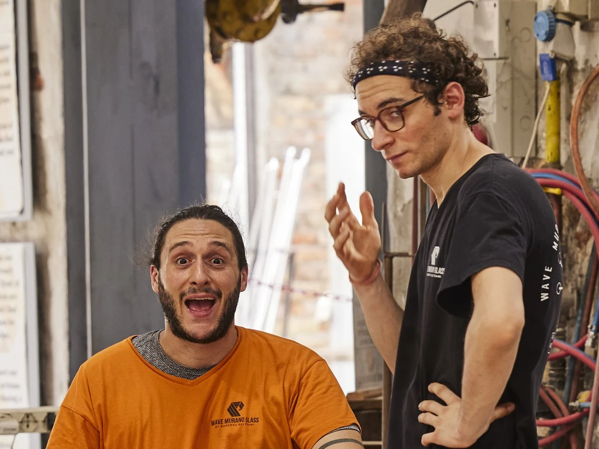 Two men in a glass workshop, one seated wearing an orange Wave Murano Glass shirt and one standing wearing a black shirt and headband, engaged in conversation.