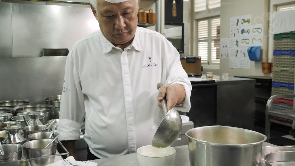 A chef in white uniform working at a counter, using a ladle to prepare a dish in a bowl, with stainless steel prep bowls visible in the background; th