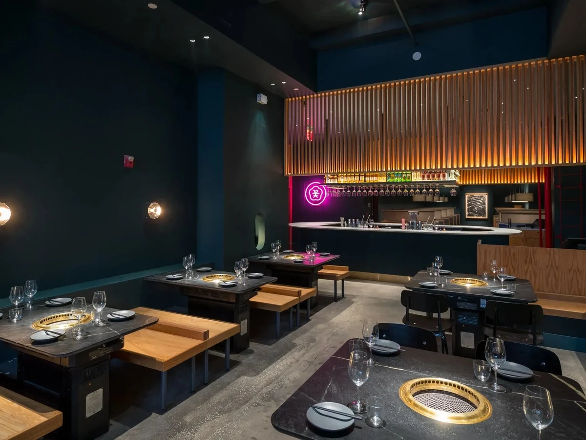 Dark-walled Korean barbecue dining room with built-in table grills, wooden bench seating, a curved bar counter, and a pink neon sign.