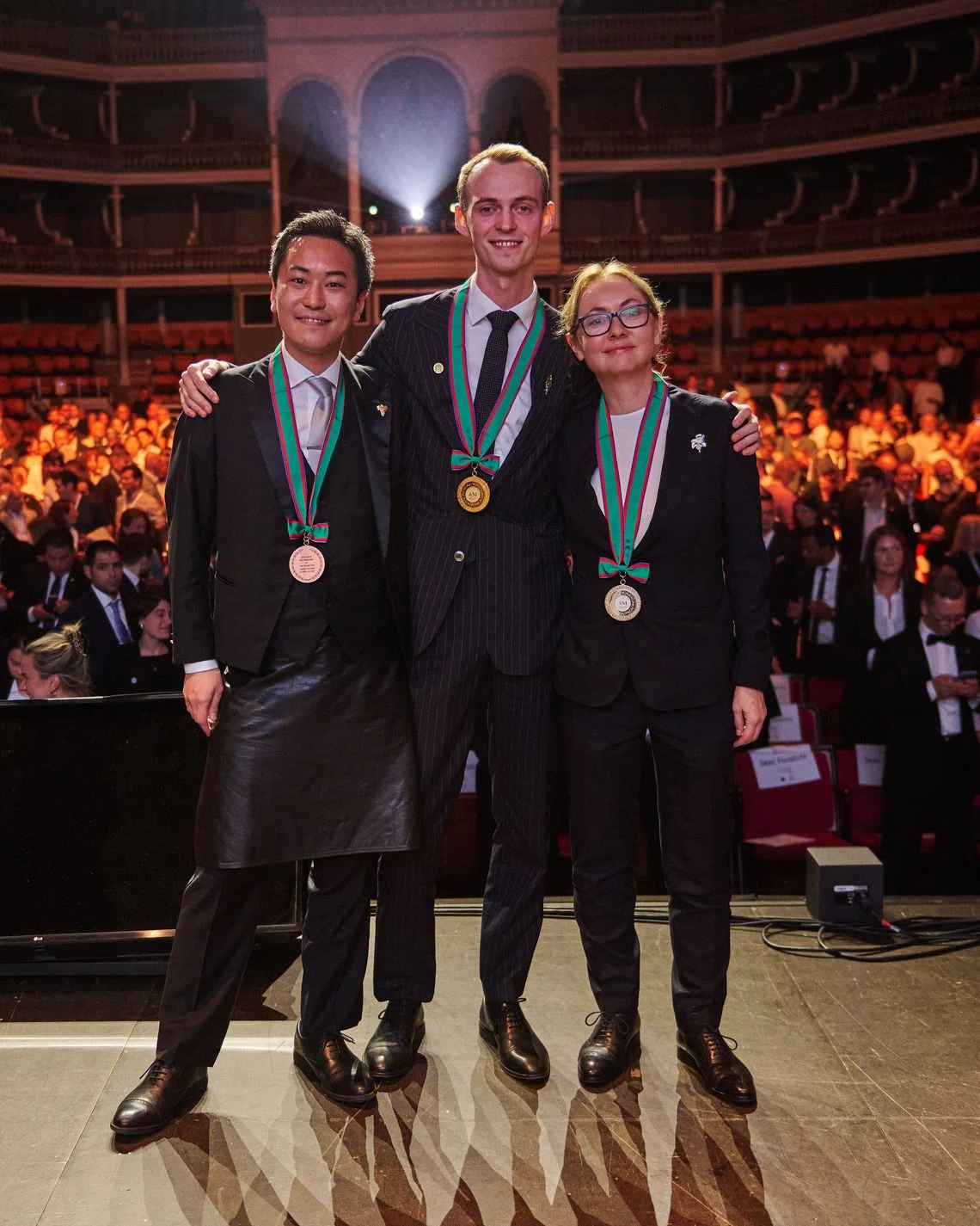 Three sommeliers in formal attire standing on stage at an awards ceremony, wearing medals (gold center, bronze flanking), in a grand theater with audi
