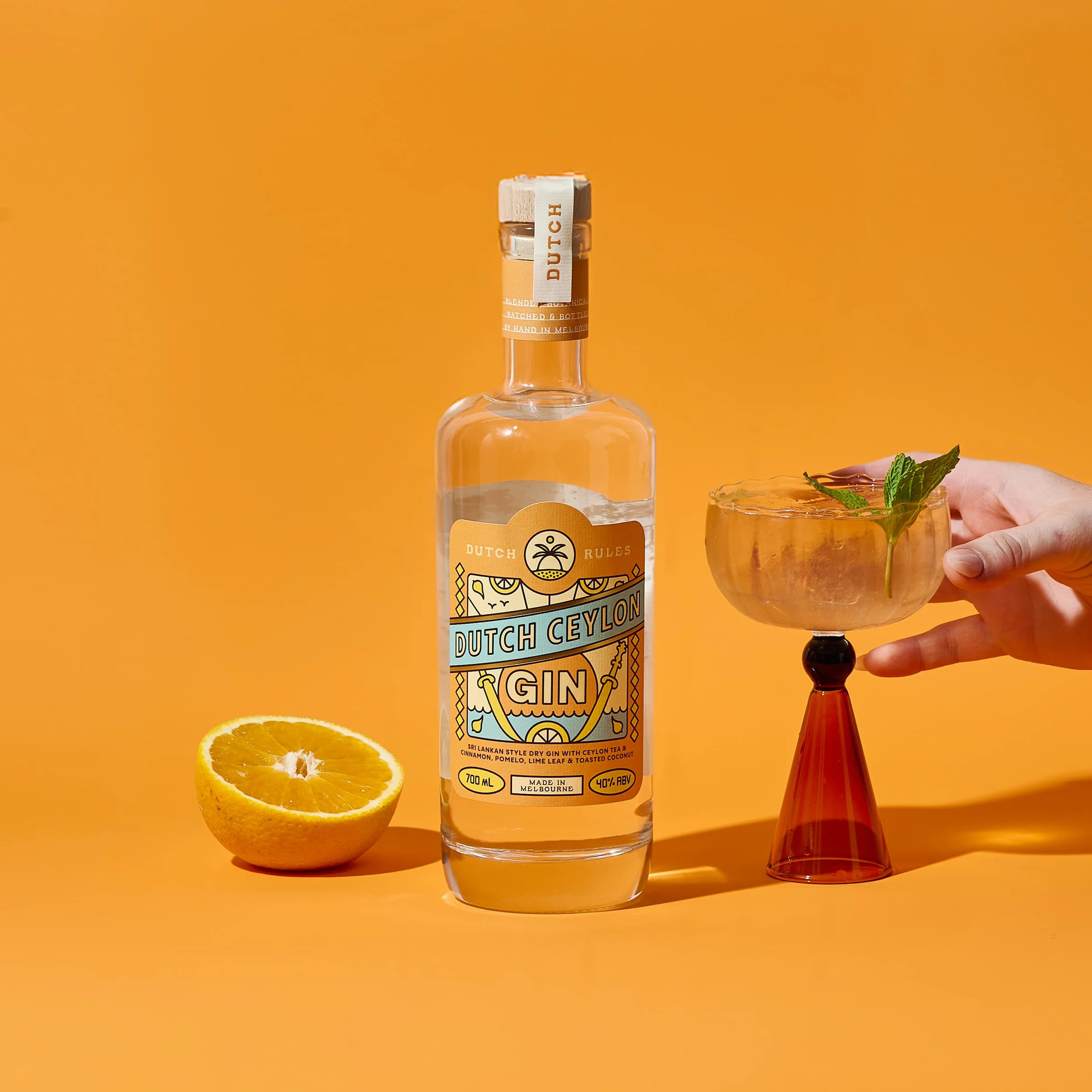 A bottle of Dutch Ceylon Gin from Dutch Rules Distilling Co. with a cocktail and lemon half on an orange background; the visible label text 'DUTCH CEY.