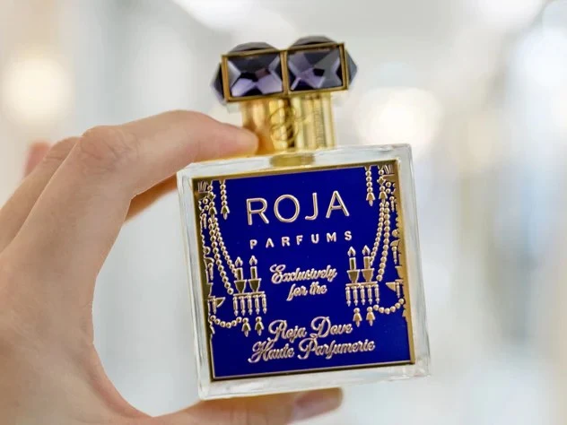 A hand holds a square Roja Parfums perfume bottle with a deep blue and gold label reading 'Roja Parfums / Exclusively for the / Roja Dove Haute